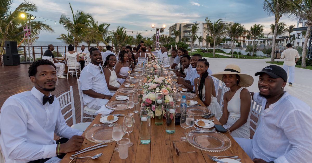 Long white destination wedding reception table set beachside with guests in white attire