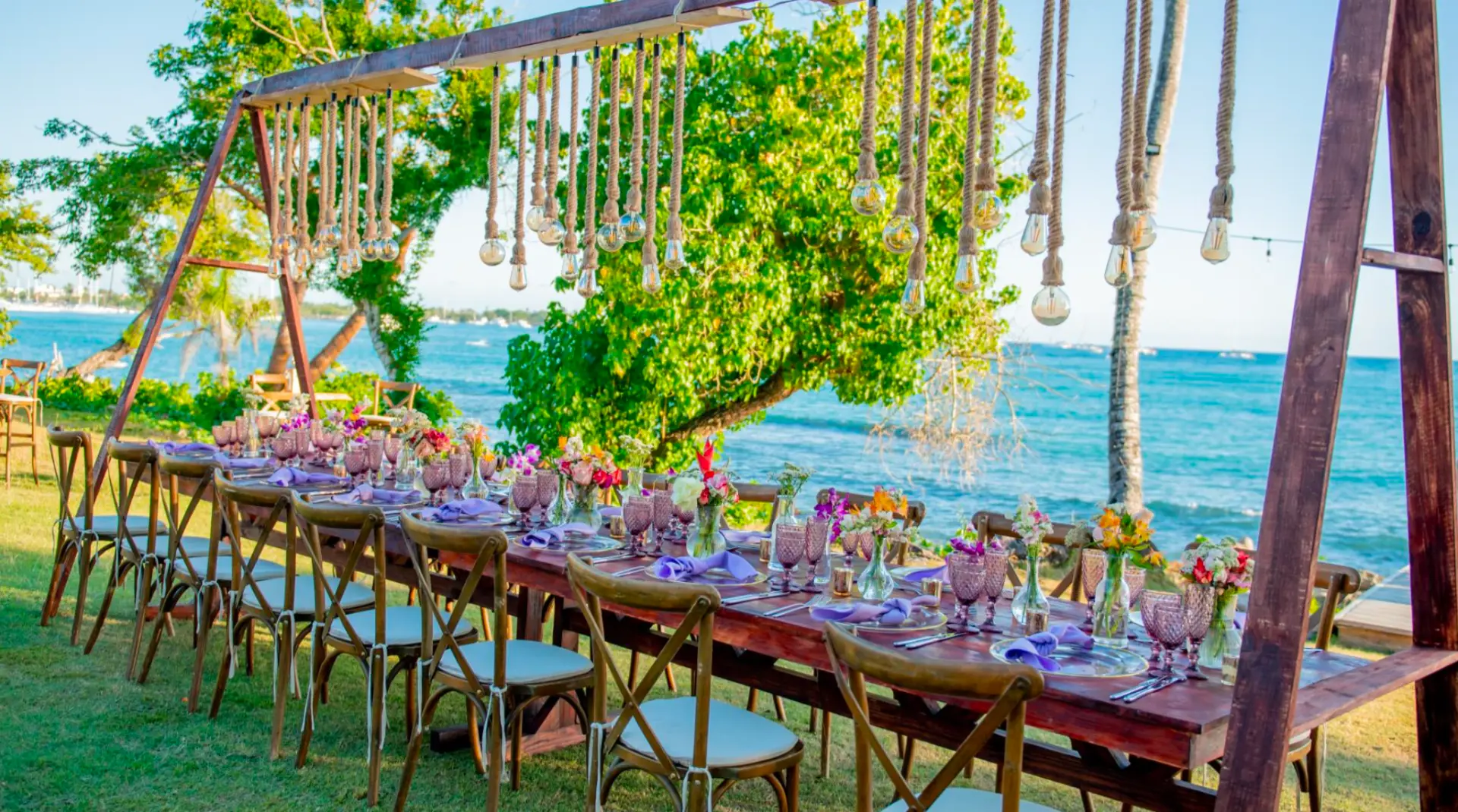 Long wooden banquet table set for a seaside garden wedding dinner on the lawn overlooking the ocean, with purple glassware