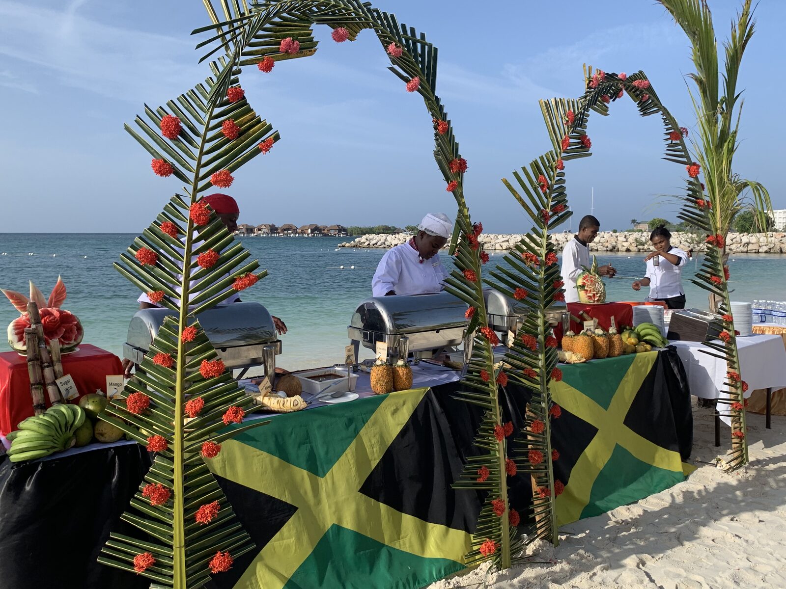 A beachfront buffet in Jamaica with the Jamaican flag flying above the food stations, the kind of local-flavor moment that carries the wedding week before the honeymoon shifts to a quieter coast