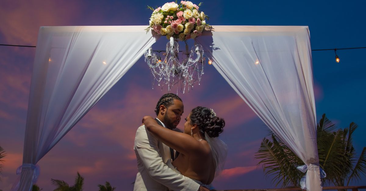 Bride and groom embrace under a draped destination wedding chuppah at sunset