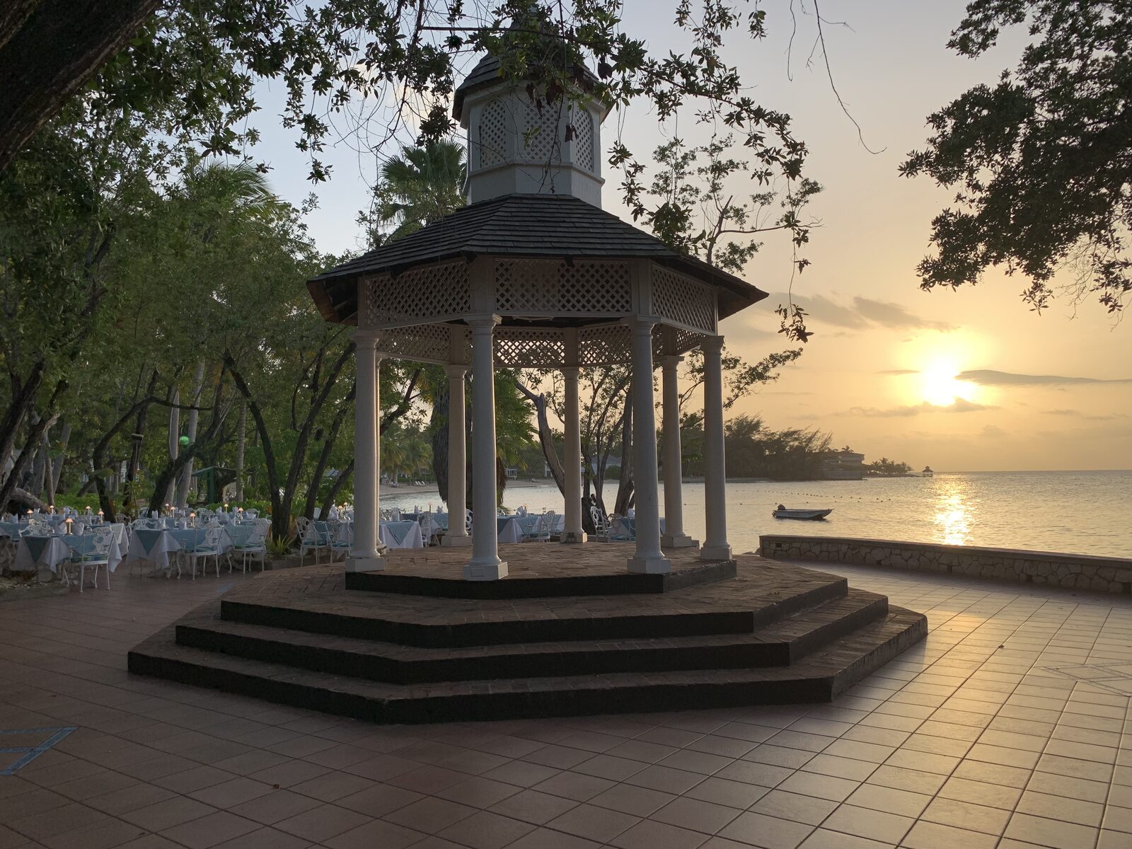 A white oceanfront gazebo at sunset with dining tables set beyond it — an example of the kind of quiet, framed spot destination wedding couples choose for reading their vows privately after the ceremony