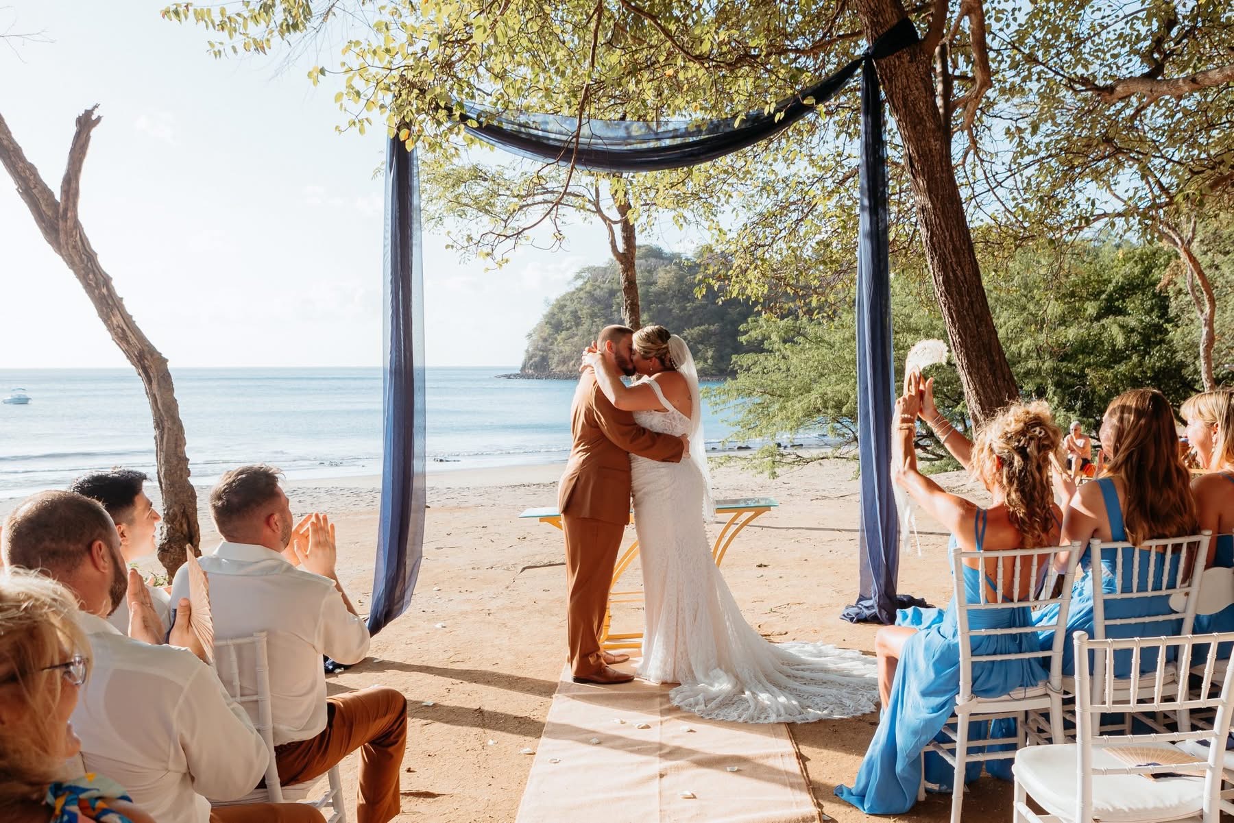 Bride and groom kiss during a beach wedding ceremony in Guanacaste, Costa Rica, with guests, ocean views, and tropical trees