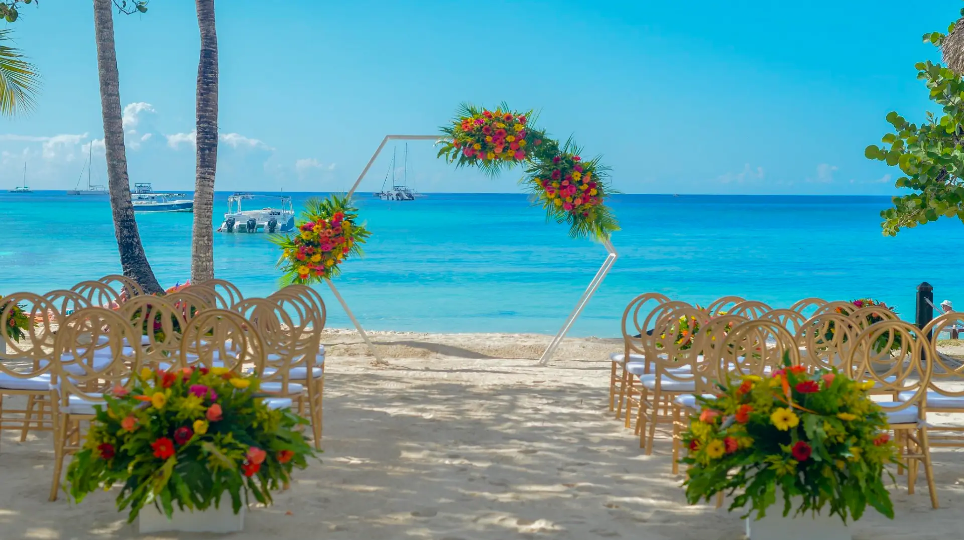 Beachfront wedding ceremony arch covered in tropical flowers on Bayahibe Beach, with turquoise water and sailboats behind it