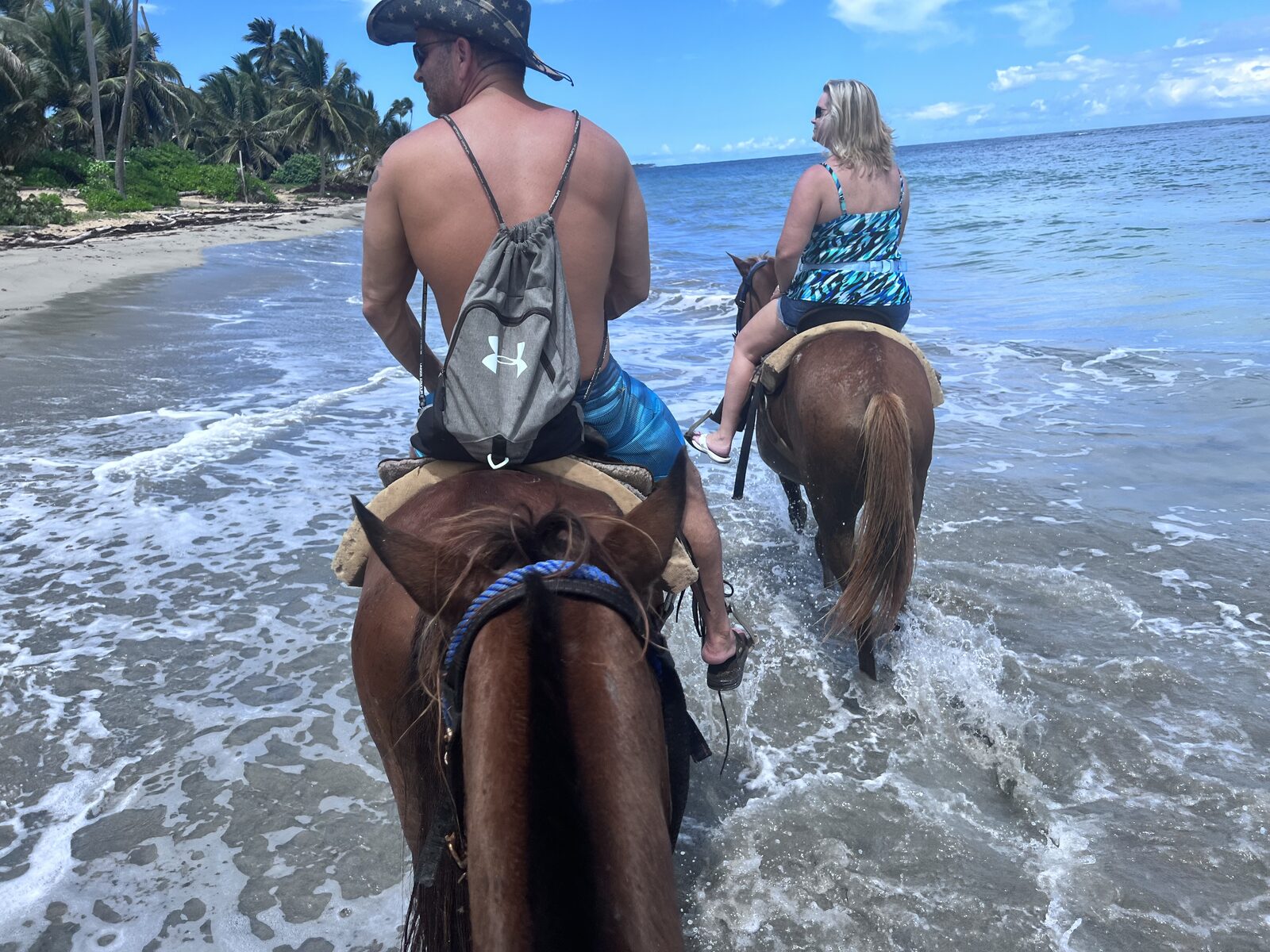 A couple horseback riding through shallow ocean water at sunset, the kind of honeymoon moment that follows a destination wedding when the wedding week and the honeymoon are planned as one trip