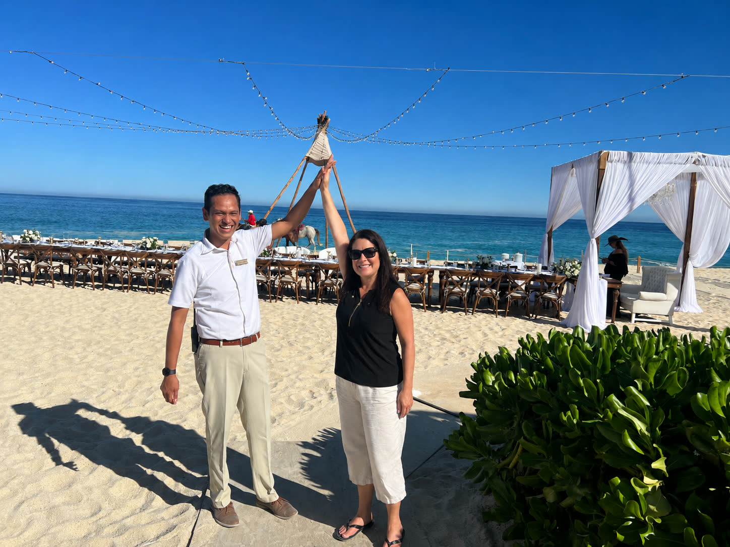 Shanna high-fiving a resort staff member on the beach in front of a styled reception setup with a draped cabana and ocean behind them