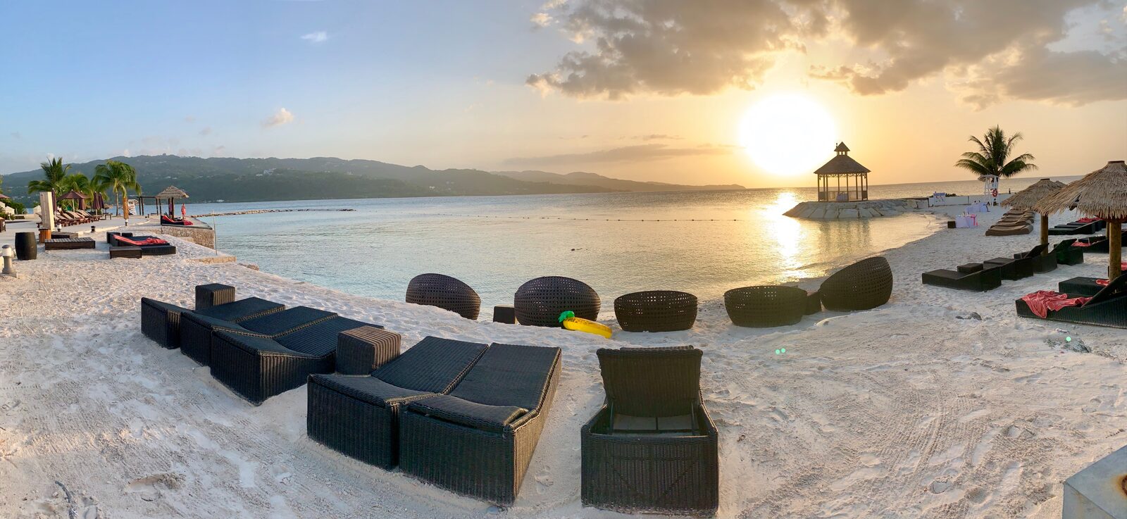 A quiet resort beach at sunset with wicker daybeds and a small oceanfront gazebo — the kind of private setting couples choose for their post-ceremony vow exchange, away from the guest crowd