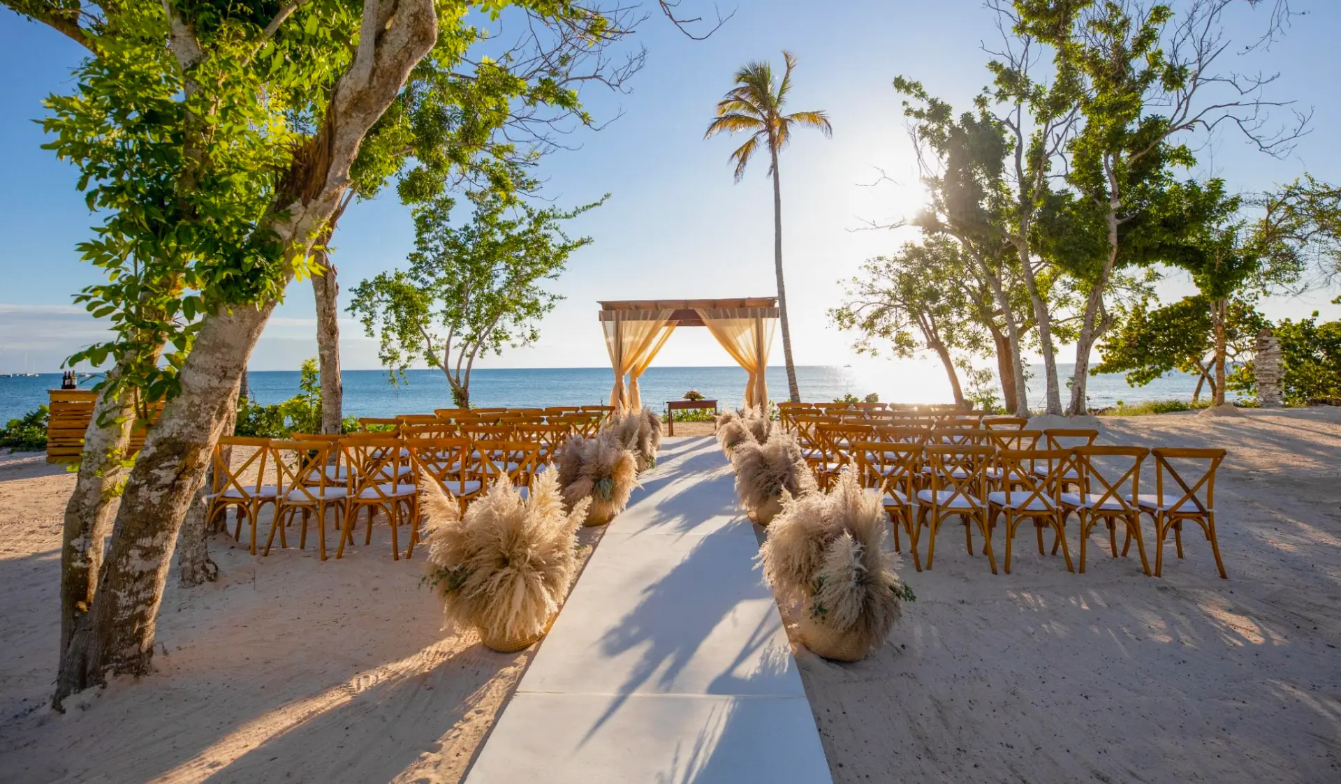Draped wooden beachfront gazebo set up for a wedding ceremony at sunset, framed by palm trees