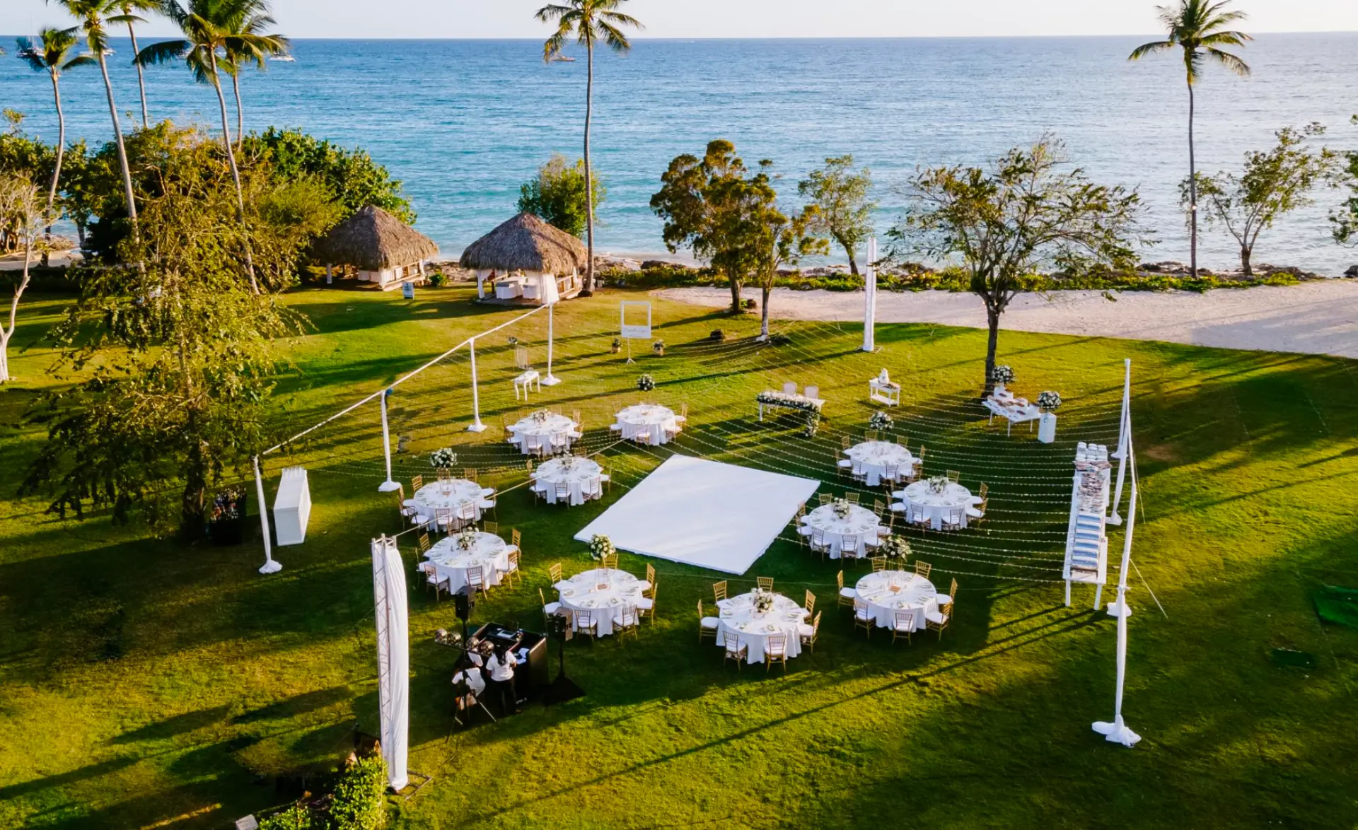Aerial view of an oceanfront Sunset Garden West reception at dusk, with round tables, a white dance floor, and crisscrossing string lights