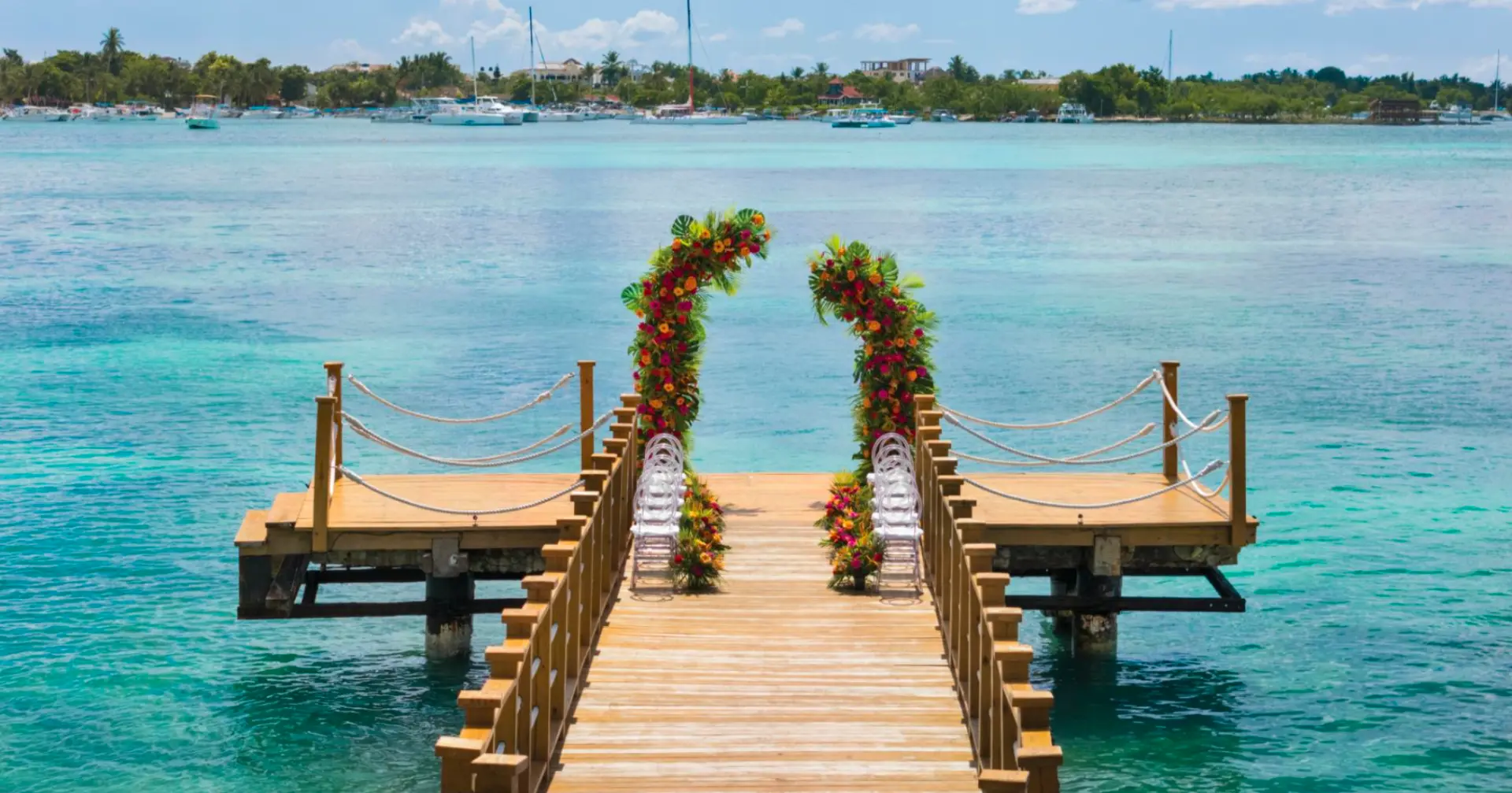 Oceanfront wooden pier wedding ceremony with tall floral pillars at the end, over turquoise water
