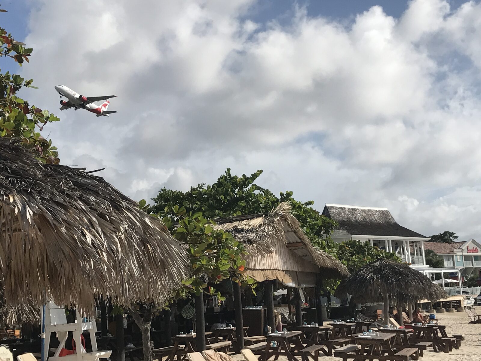 A commercial airplane passing low over a resort beach with thatched palapas and picnic tables — the exact moment where a well-chosen airport transfer takes travel stress off the plate and lets a wedding trip actually start well