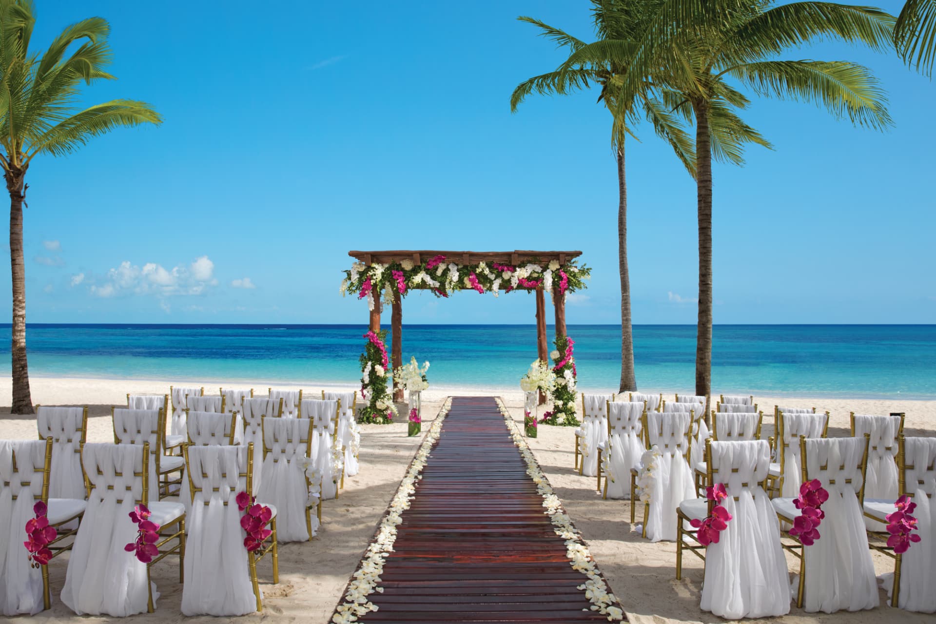 Oceanfront beach wedding ceremony aisle with a floral gazebo, white chiavari chairs, and palm trees.