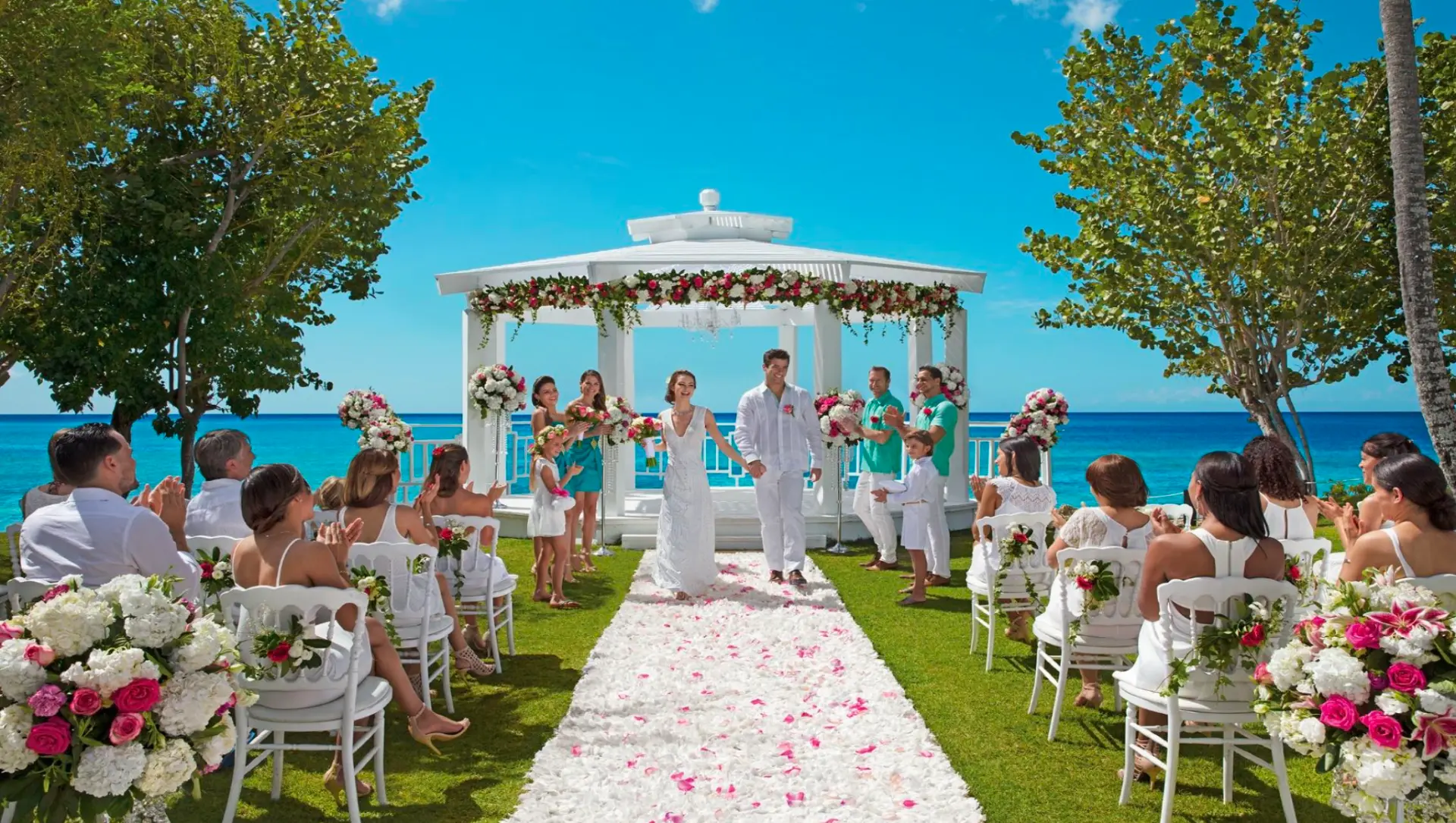 White octagonal garden gazebo wedding ceremony by the sea, with the couple at the altar and guests seated in white chairs