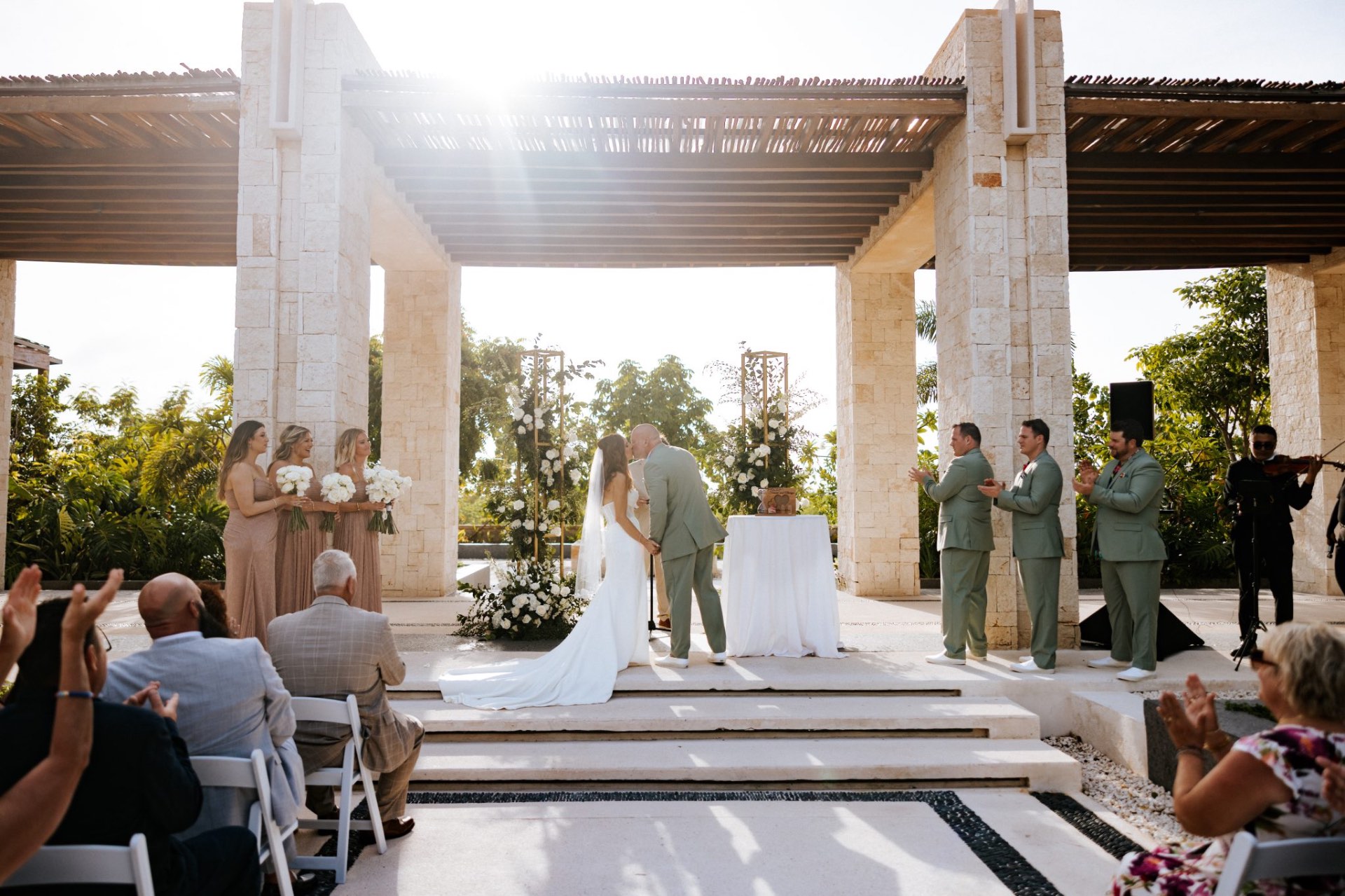 Bride and groom sharing their first kiss as newlyweds under a stone pergola ceremony arch at Secrets Playa Blanca, surrounded by bridesmaids, groomsmen, and applauding guests