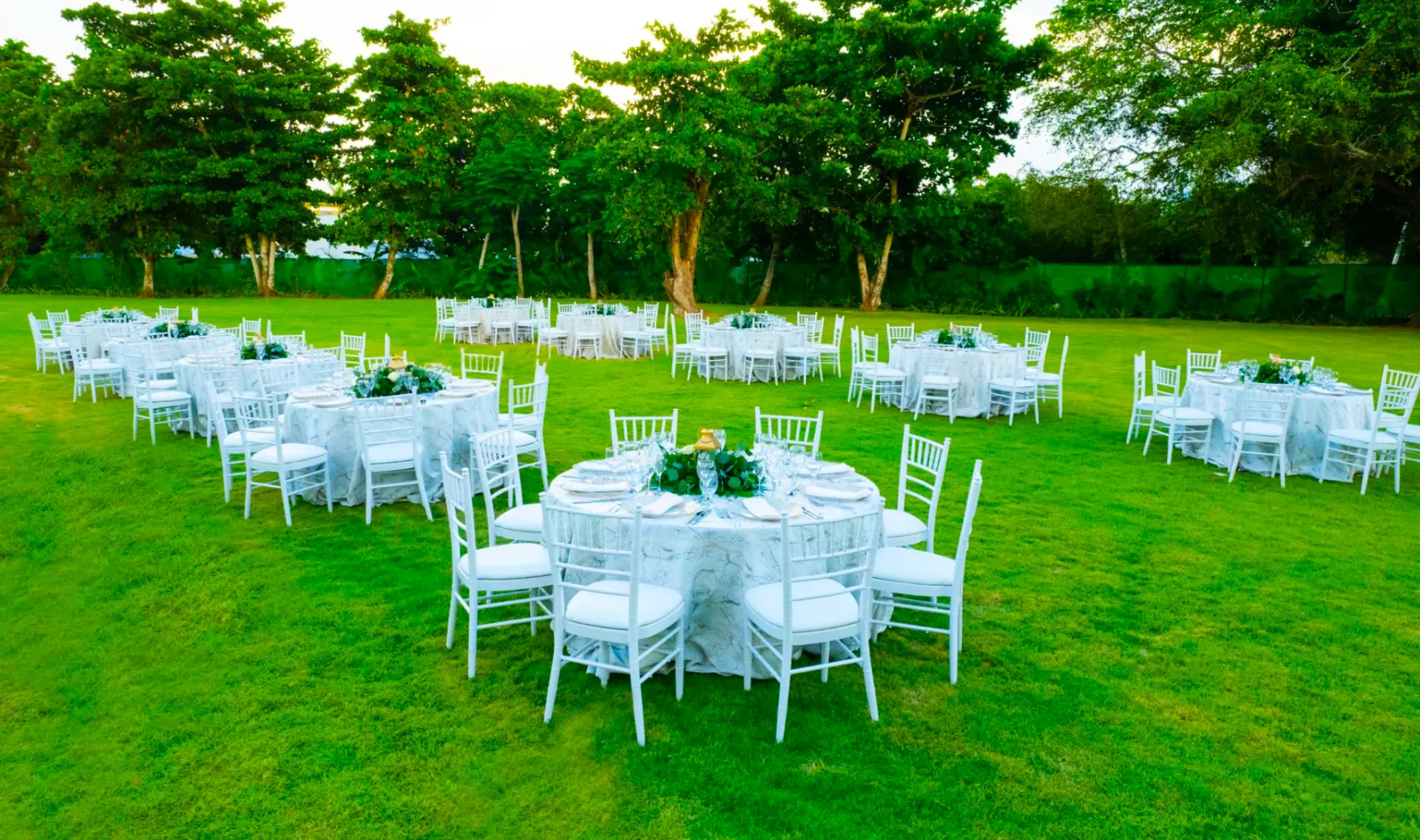 Coco Garden wedding reception with round tables in white linens and white chairs on a wide green lawn ringed by trees